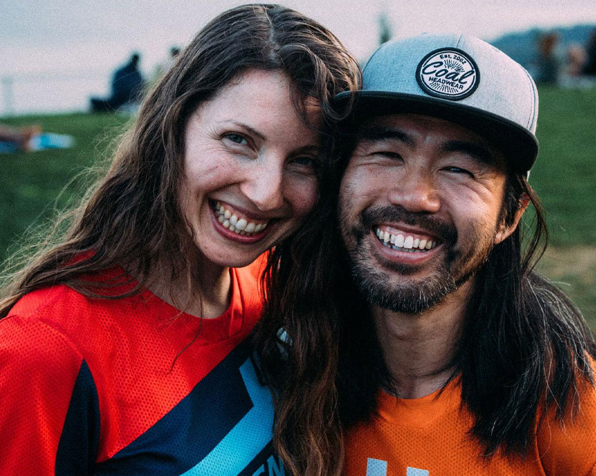 Two people smiling at the camera with a grassy field and people in the background.